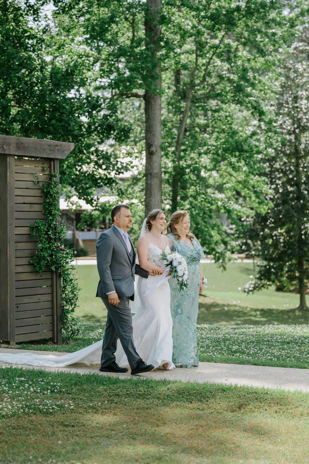 Bride walks down outdoor aisle with parents on sunny day, holding white bouquet, wearing elegant white gown.