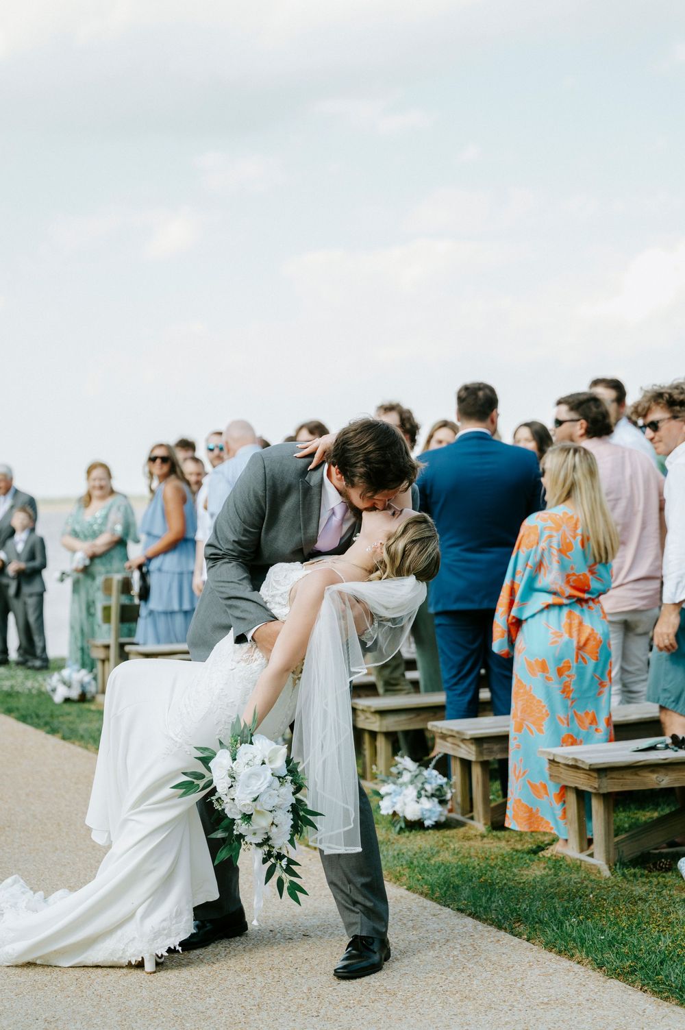 Groom dips bride for a romantic kiss at outdoor wedding ceremony while guests celebrate in the background.