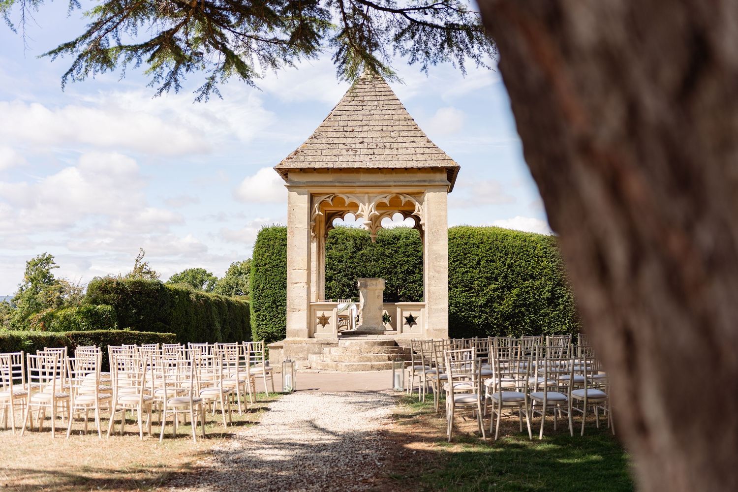 Elegant outdoor wedding ceremony setup with white chairs and a stone gazebo surrounded by manicured hedges.