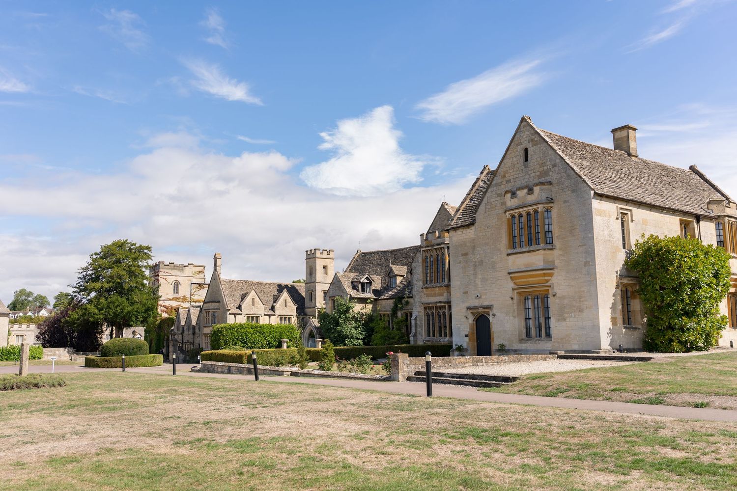Historic Cotswold stone manor house with church tower under blue sky, surrounded by manicured lawns and mature gardens.