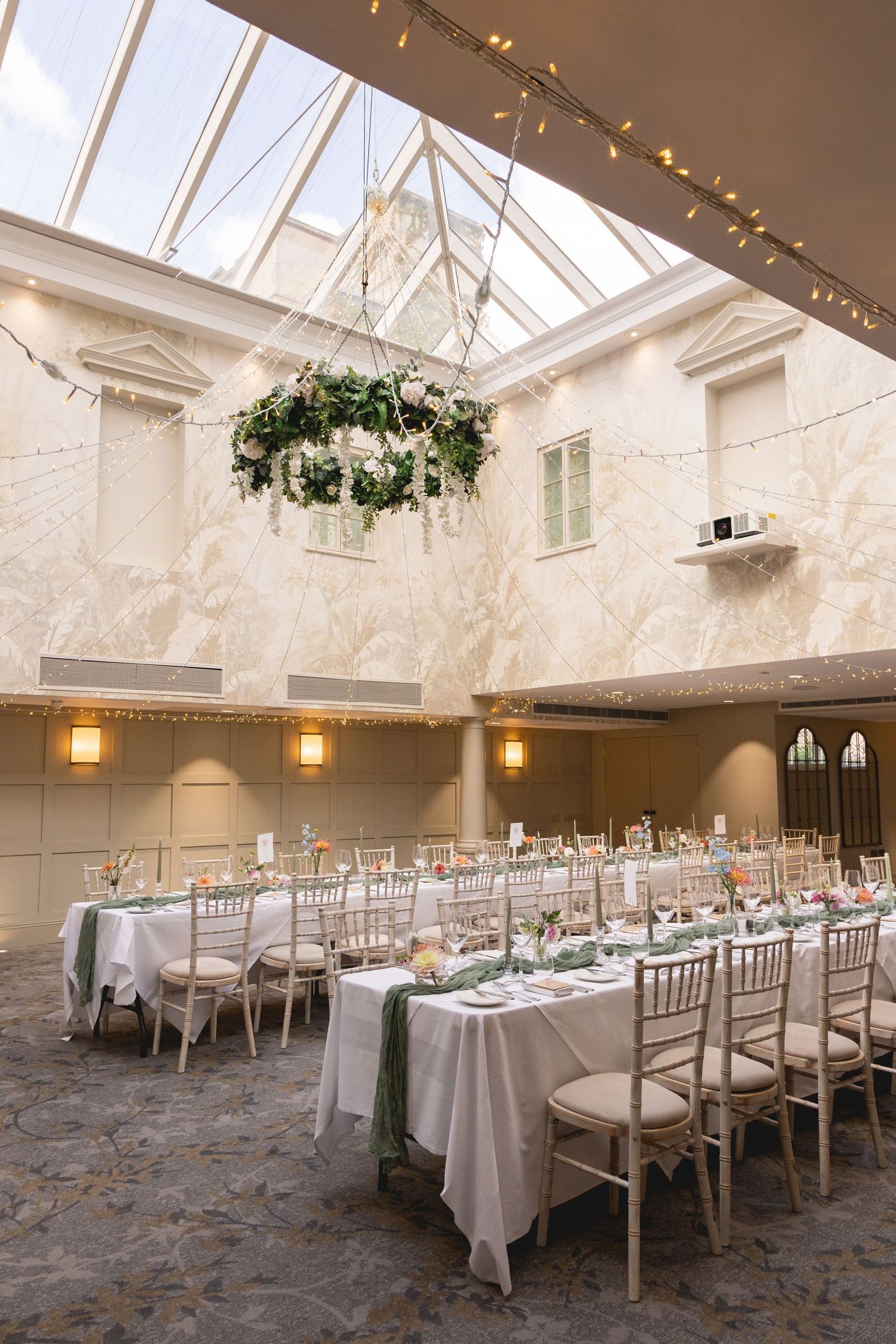 Elegant wedding reception hall with white tablecloths, chiavari chairs, floral chandelier, and glass skylight ceiling.