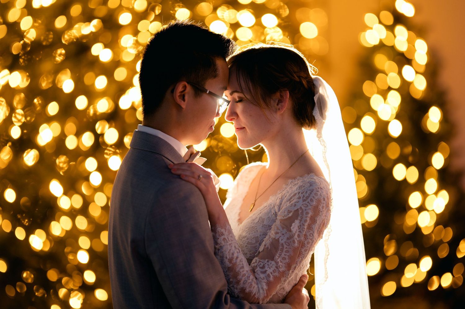 Couple sharing romantic moment in front of glowing Christmas tree lights, bride in lace veil and groom in suit.
