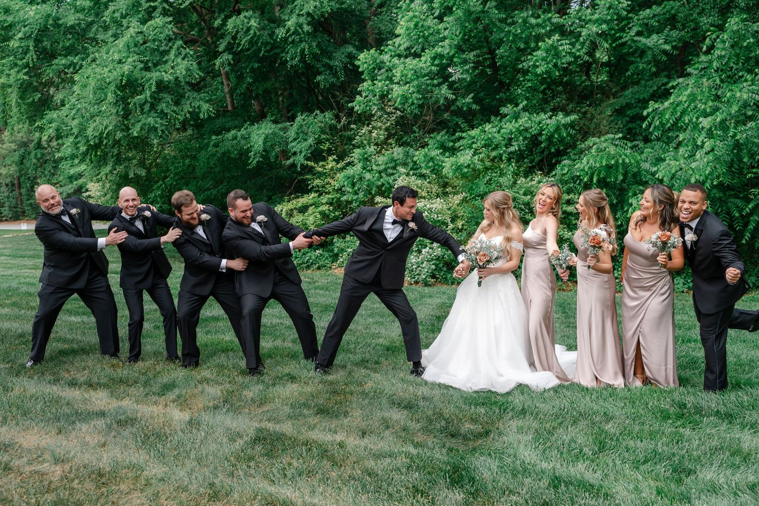 Wedding party poses humorously with groomsmen pulling groom toward bridesmaids on lush green lawn.