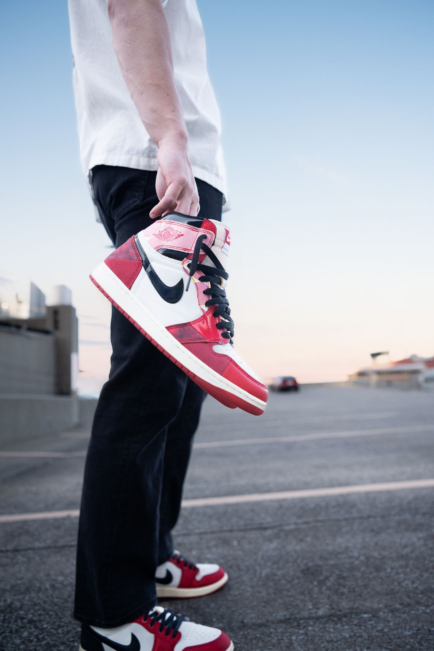 Person holding red and white Nike Air Jordan 1 High sneakers outdoors at dusk on a rooftop parking lot.