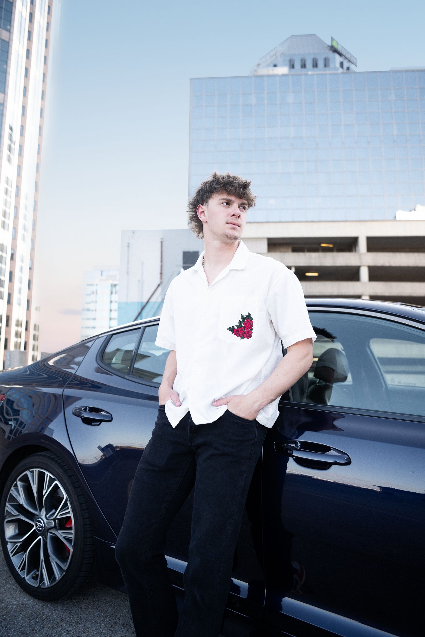 Young man in white floral shirt and black jeans leaning on dark luxury car with city buildings in background.