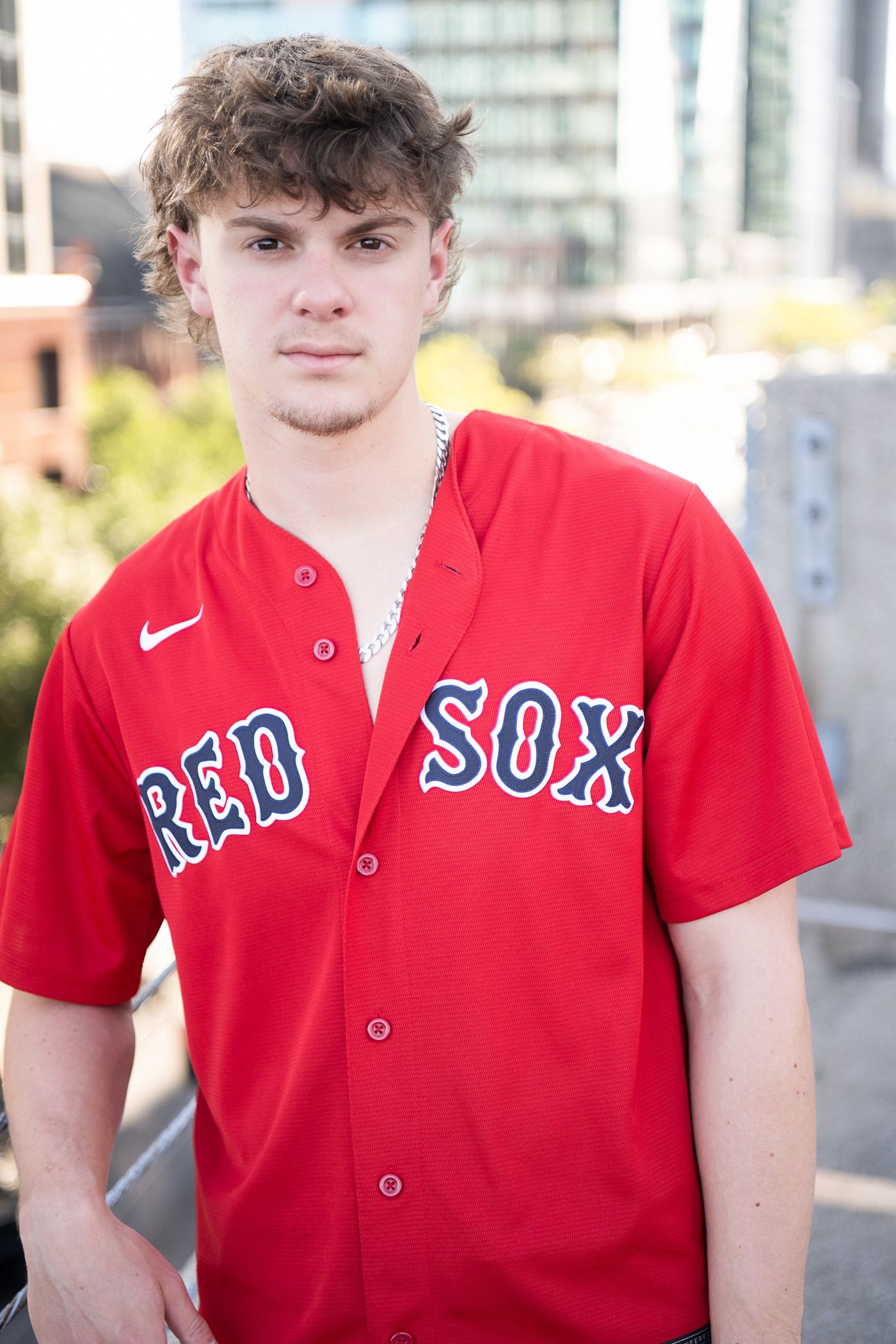 Young man wearing a red Nike Boston Red Sox baseball jersey outdoors with urban buildings in the background.