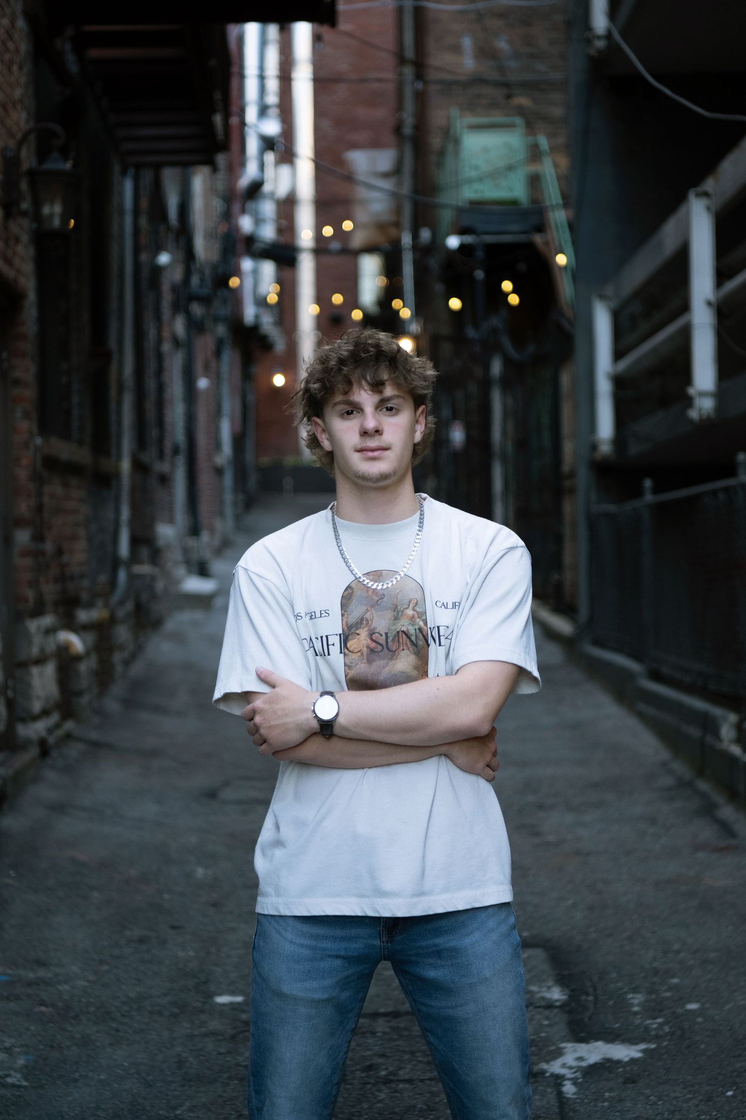 Young man with curly hair stands arms crossed in urban alley wearing white graphic tee, jeans, and watch.