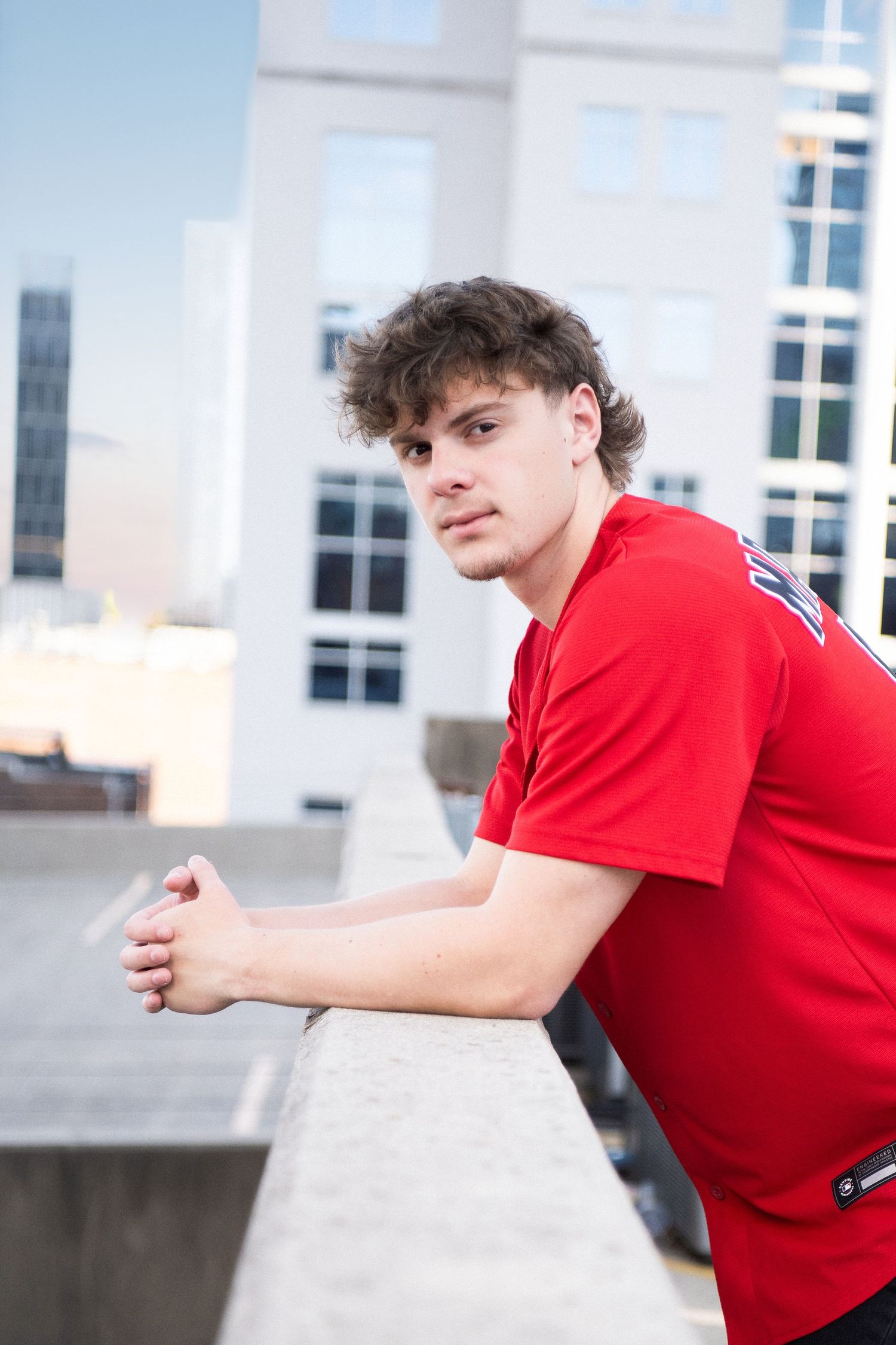 Young man in red t-shirt leaning on rooftop ledge with urban city buildings in background.