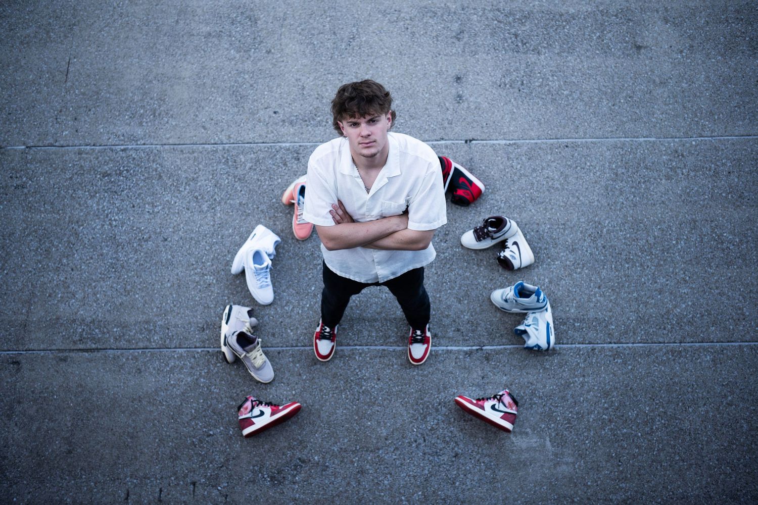 Young man sitting cross-legged on pavement surrounded by Nike Air Jordan sneakers, arms crossed, wearing white shirt.