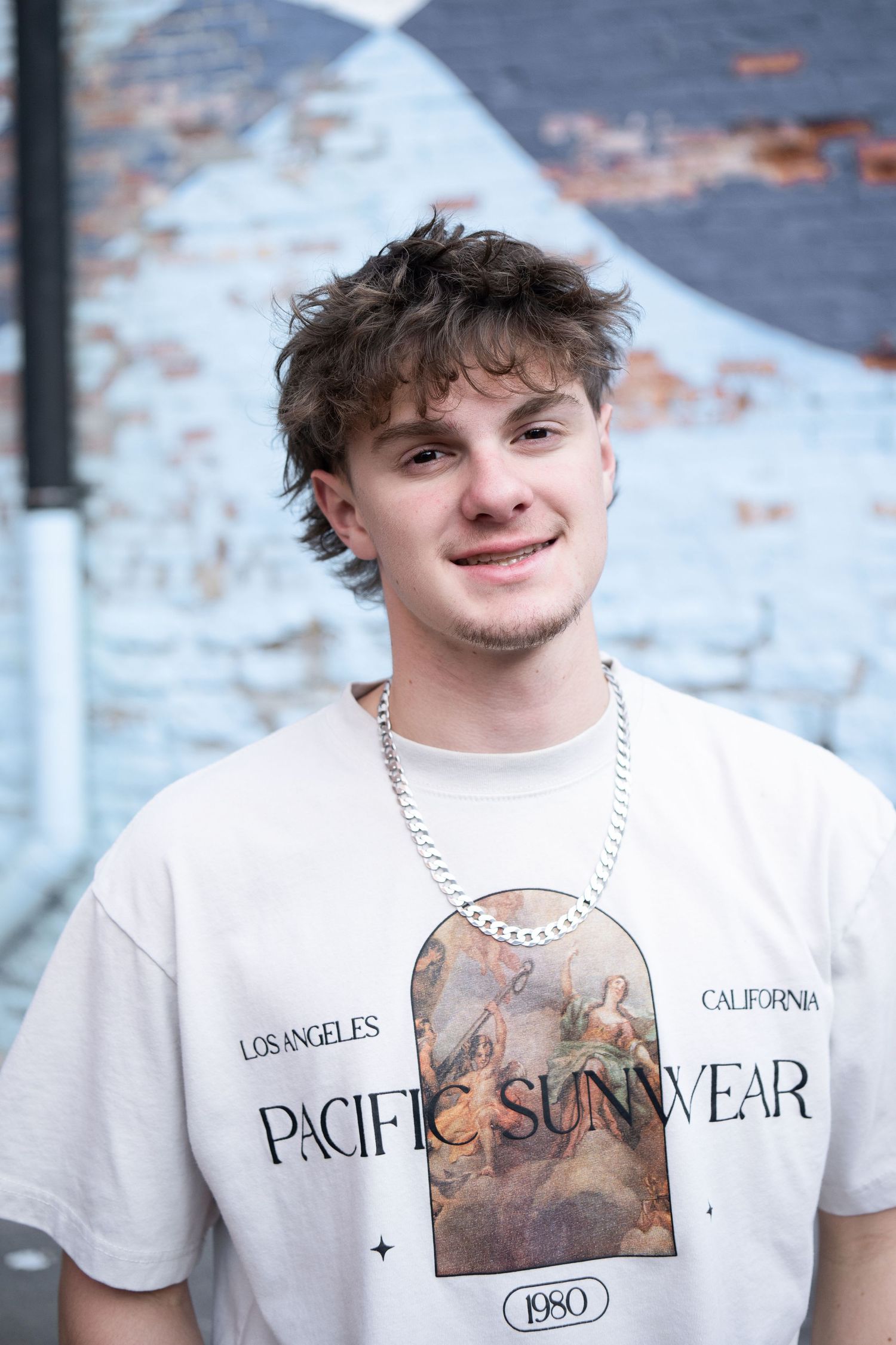 Young man in Pacific Sunwear tee and silver chain smiles outdoors against a painted brick wall.