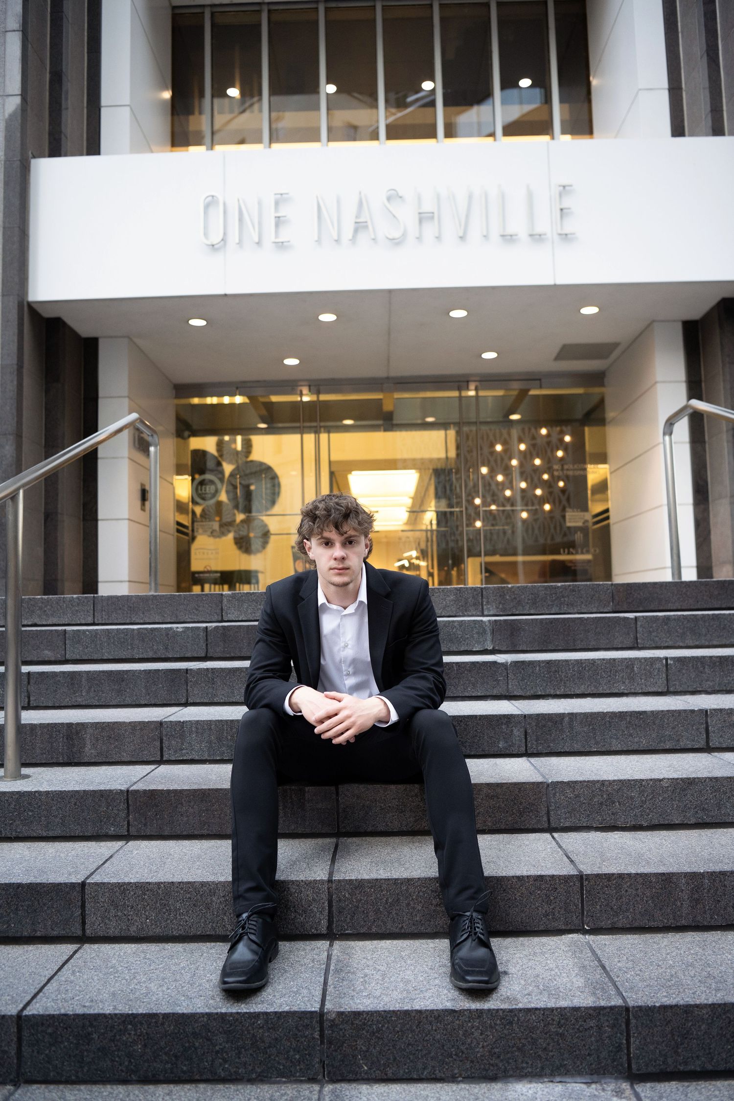Young man in black suit sitting on steps outside One Nashville building entrance.