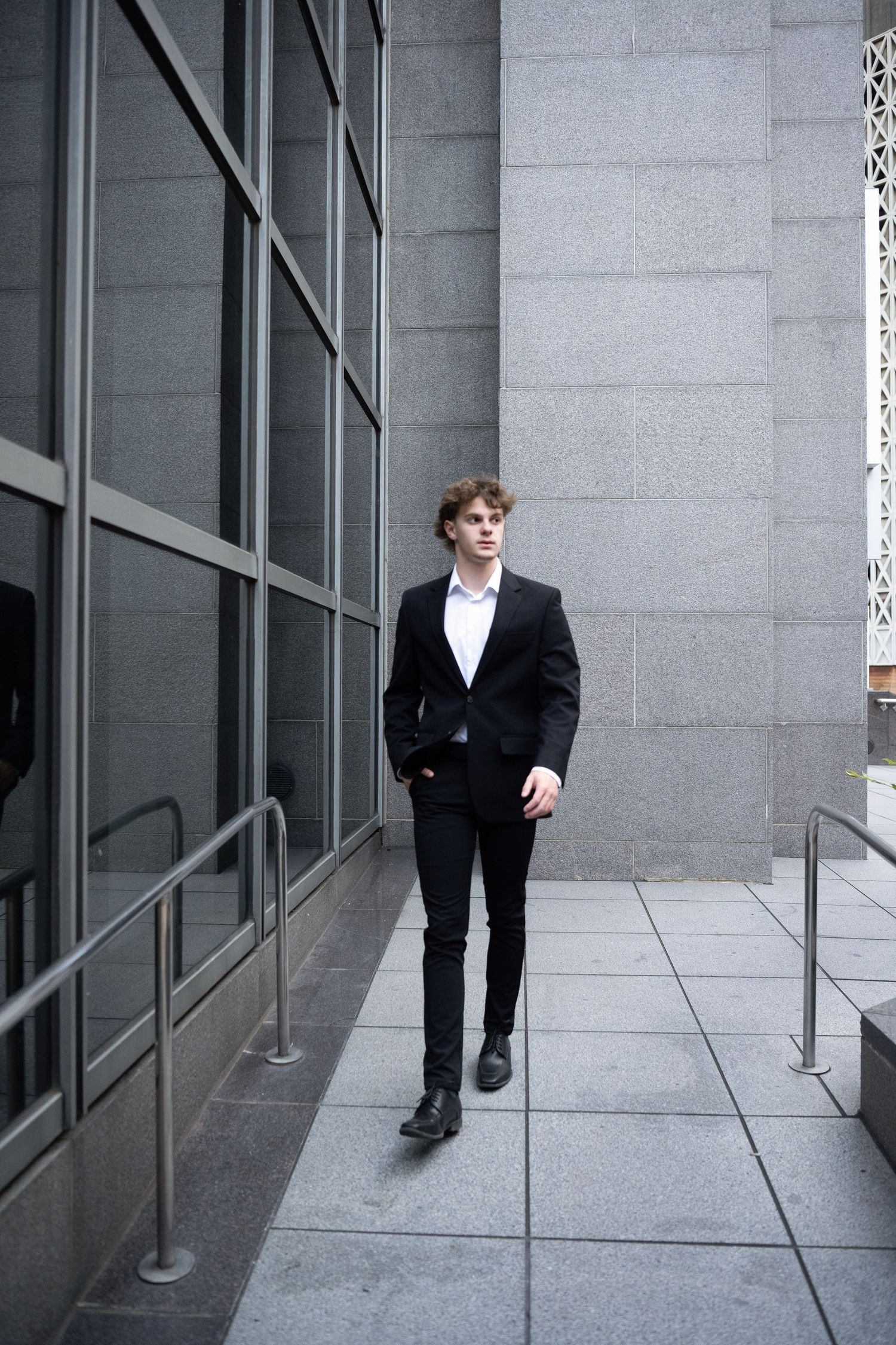 Young man in black suit walks confidently outside modern gray stone office building.