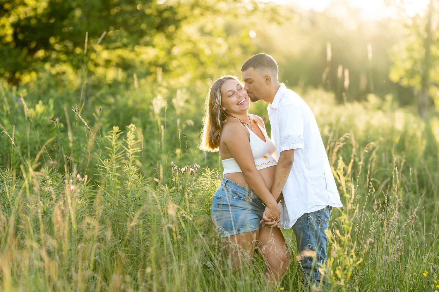 A couple embraces and holds hands in a sunlit green meadow, the man kissing the smiling woman's cheek.
