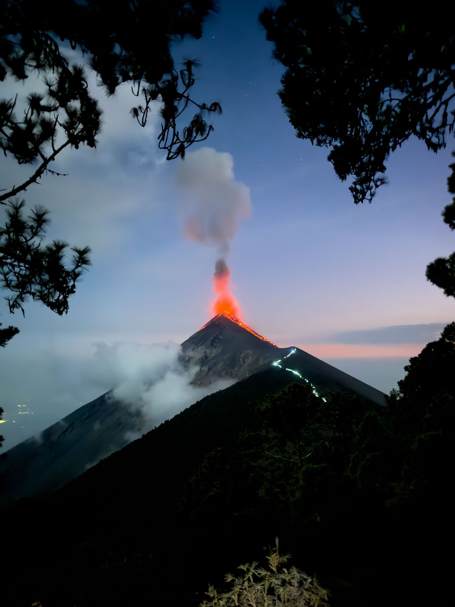 Volcanic eruption at sunset shows glowing lava and smoke against darkening sky through silhouetted trees.