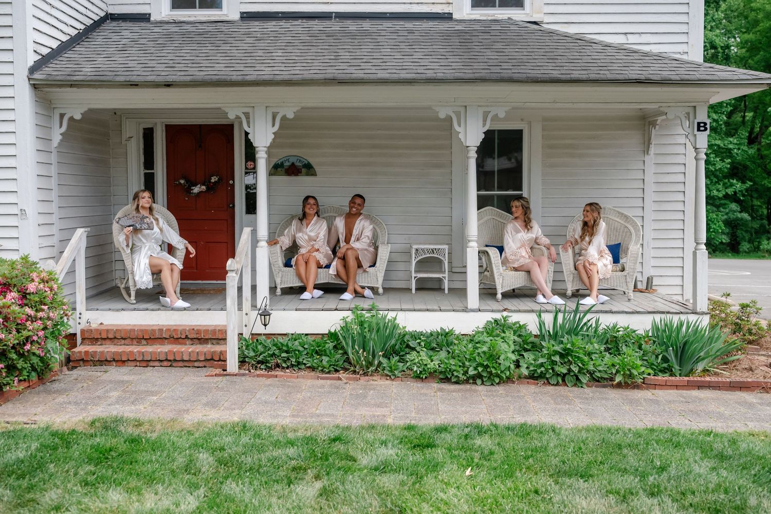 Women in matching robes relaxing on a white farmhouse porch with red door, lush greenery, and brick steps.