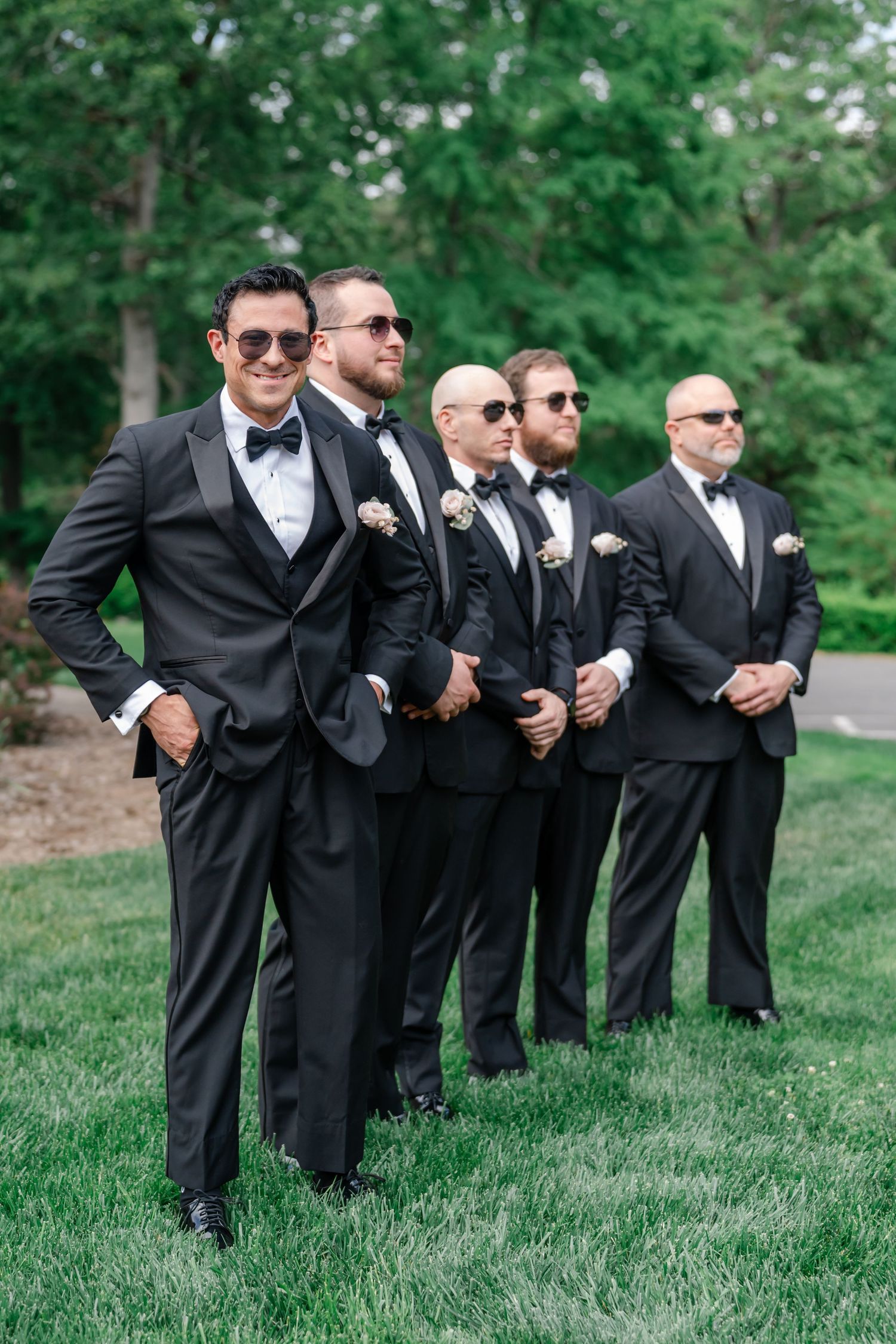 Five groomsmen in black tuxedos and sunglasses pose on green grass at an outdoor wedding ceremony.