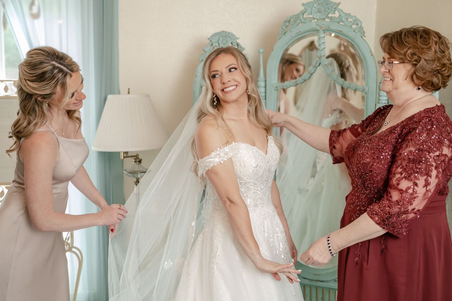 Bride in white gown and veil smiles as bridesmaid and mother help her prepare in elegant room.