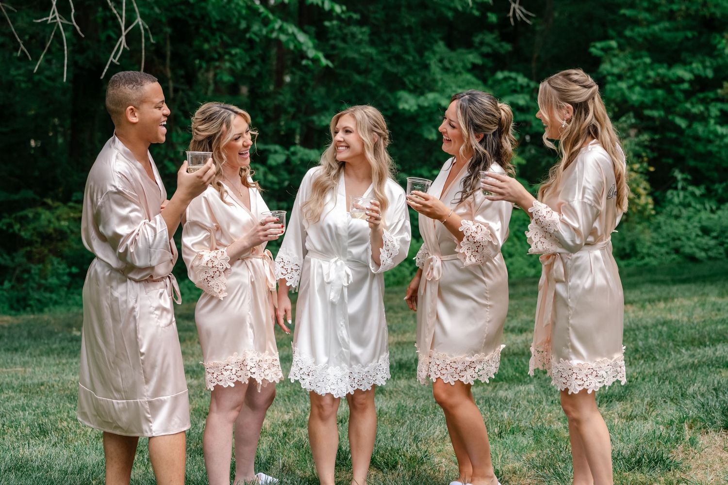 Five bridesmaids in matching champagne satin robes toasting with glasses outdoors among lush green trees.