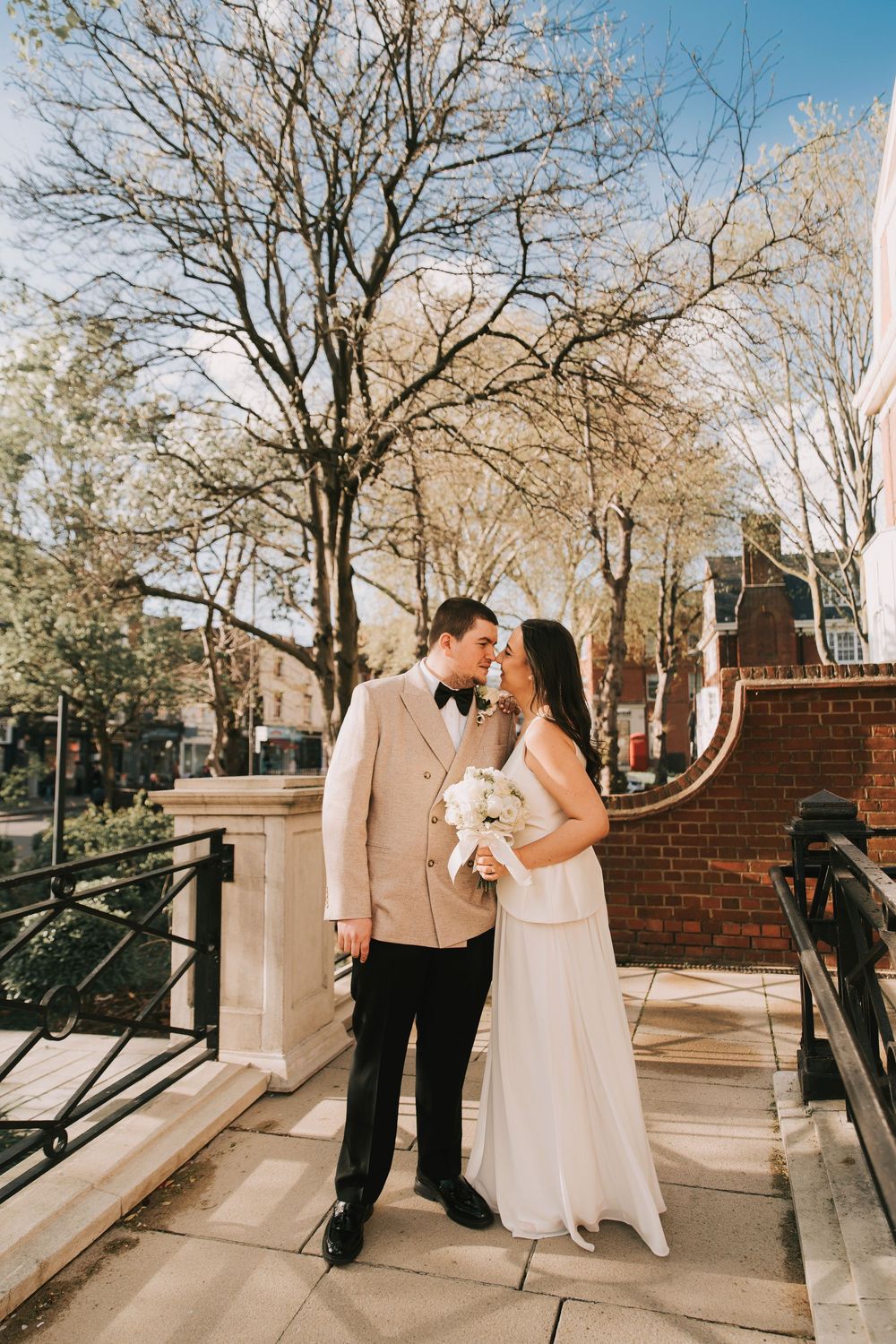 Bride and groom share a romantic moment outdoors, she holds white bouquet, he wears tan tuxedo jacket with black bow tie.