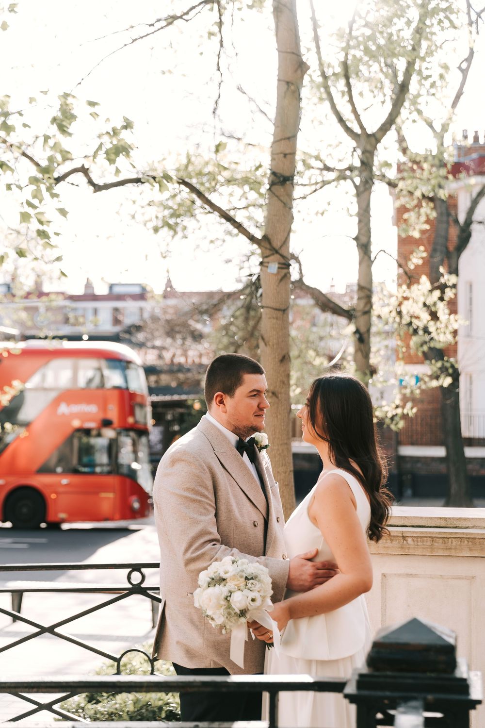 Bride and groom holding white bouquet with red London double-decker bus in background on sunny wedding day.