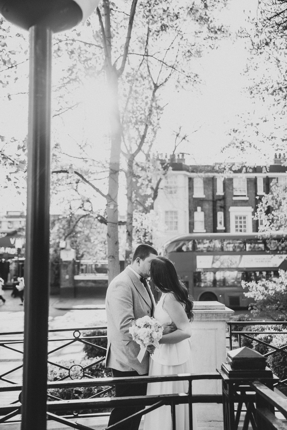 Black and white photo of a couple kissing on a London street, bride holding bouquet with a double-decker bus in background.