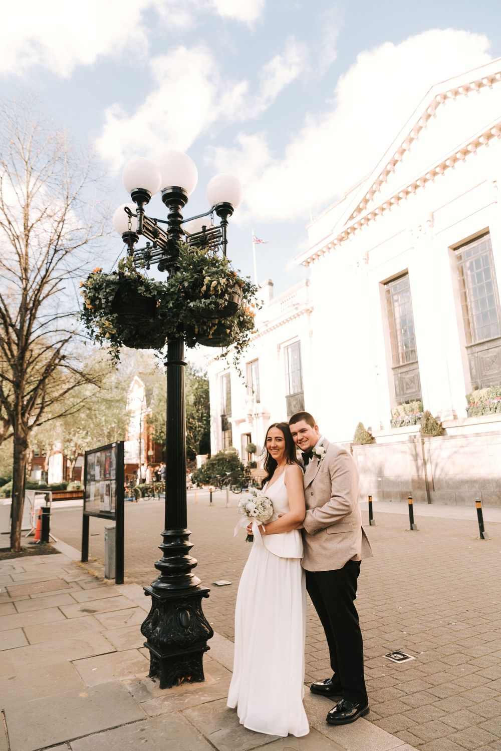 A bride and groom pose together by an ornate street lamp outside a white building on their wedding day.