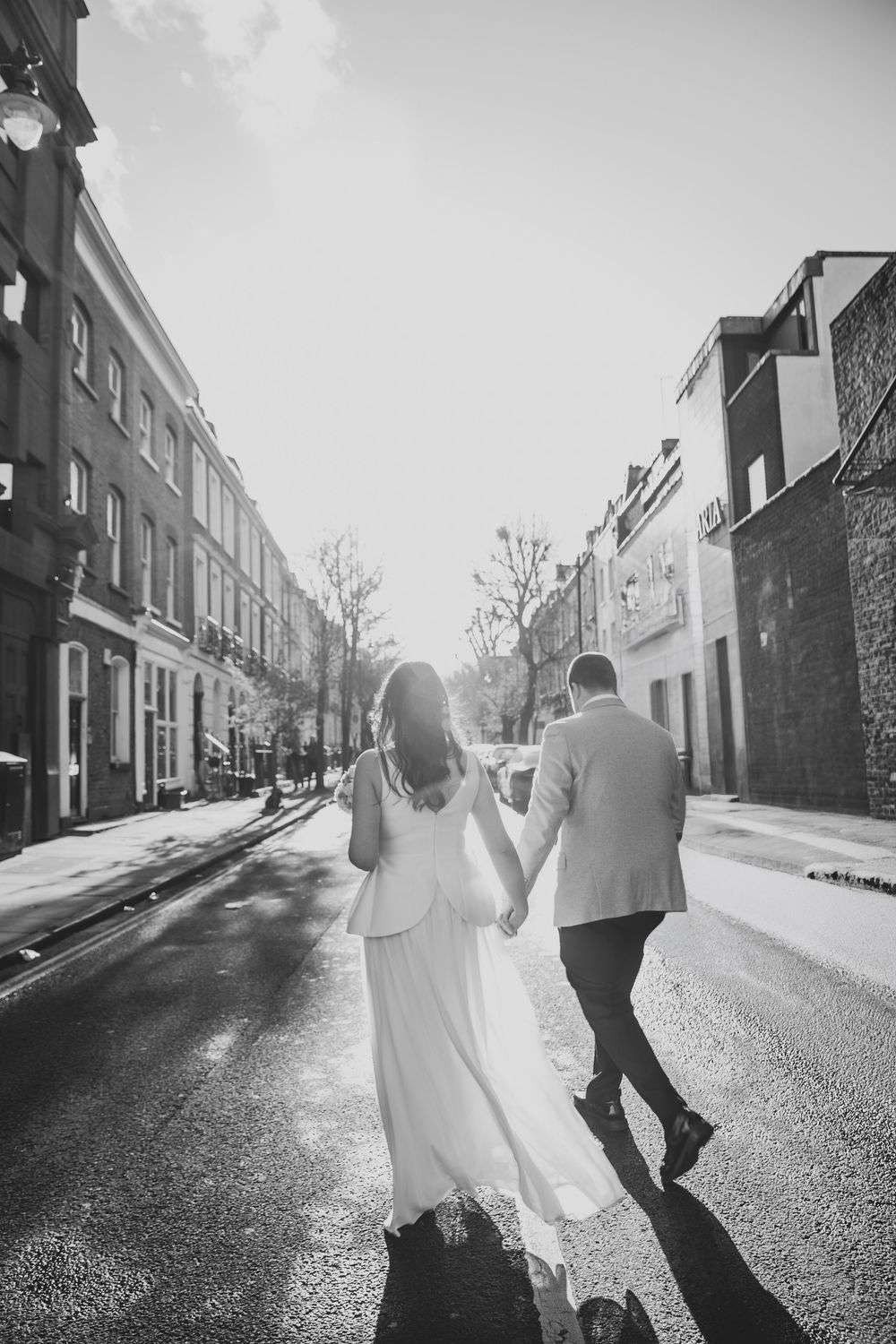 Black and white photo of bride and groom walking hand in hand down a sunlit London street.