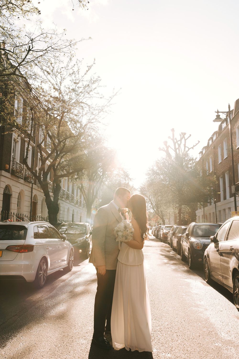 Couple sharing a romantic kiss on a sunlit London street, bride in white gown holding bouquet, cars parked alongside.