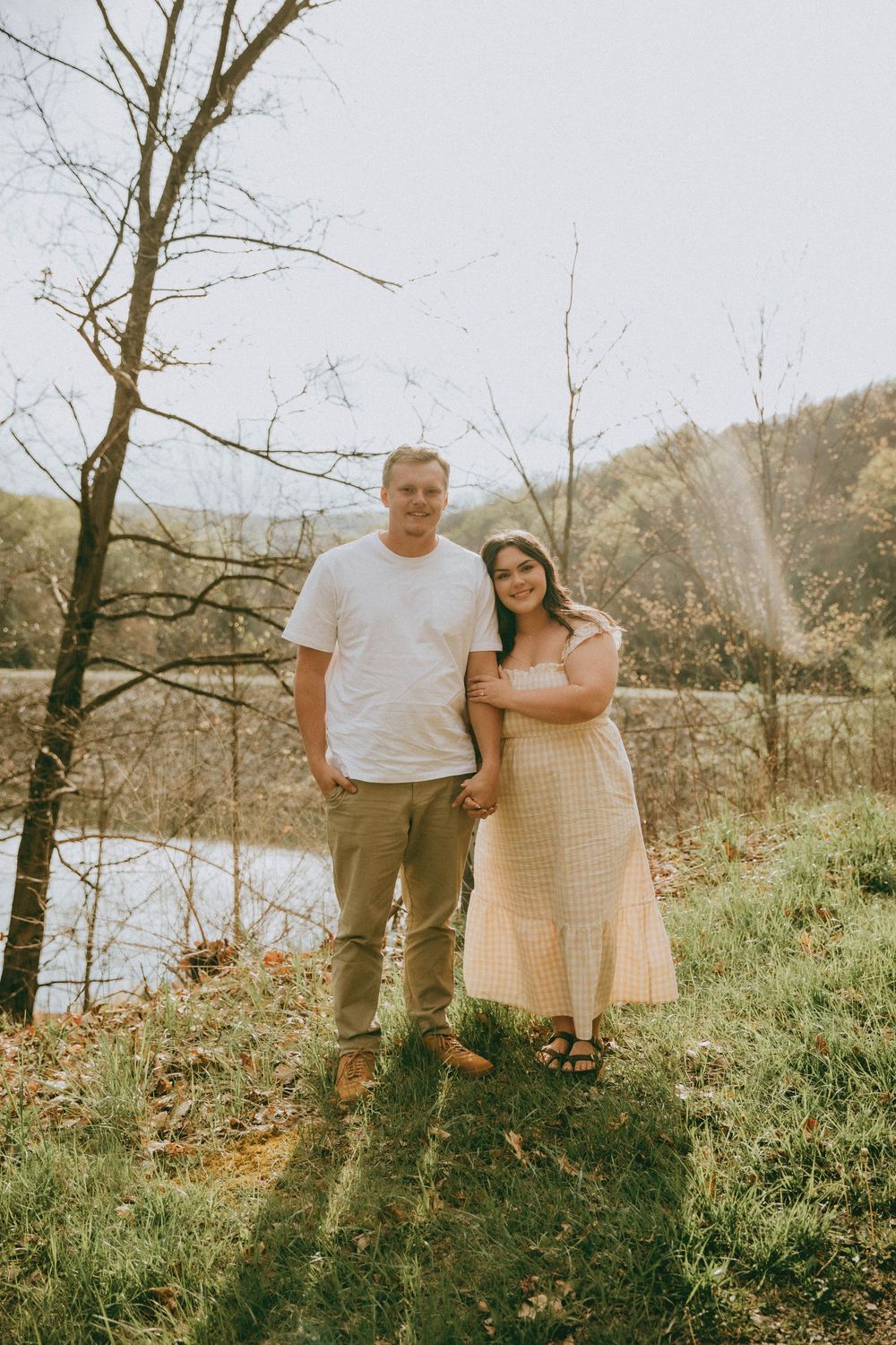 Couple posing outdoors near a pond in golden hour spring, man in white tee and khakis, woman in yellow floral midi dress. engagement session