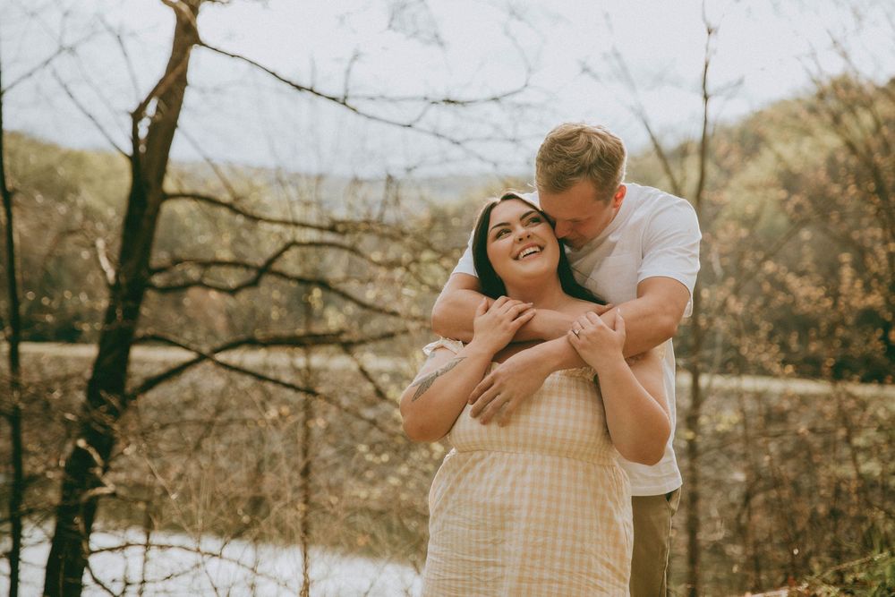 Couple embracing outdoors in spring, man kissing woman's cheek as she smiles, trees and river in background. indiana pa photographer