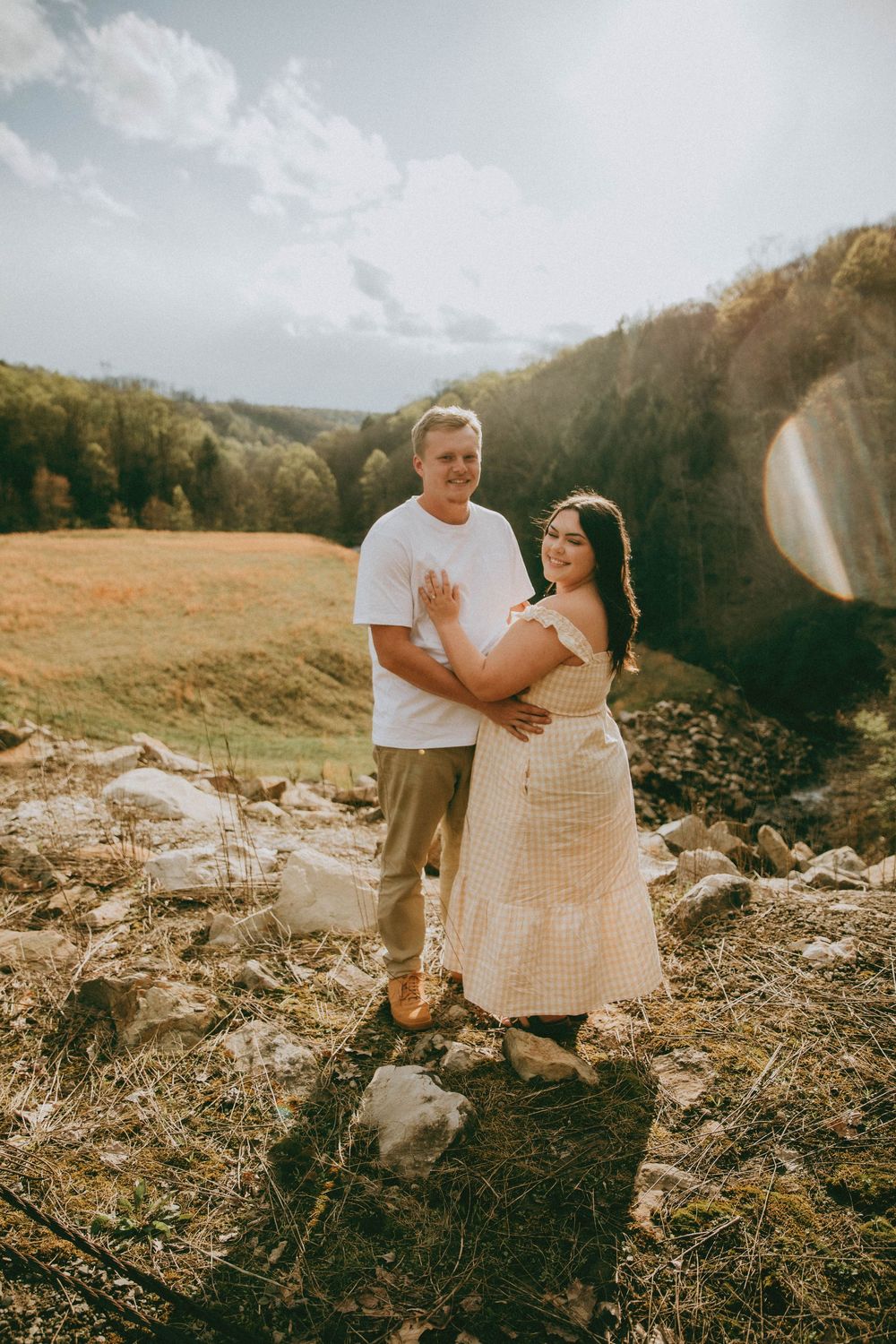 A couple poses outdoors on rocky terrain, woman in floral dress with baby bump, man in white tee, scenic valley. indiana pa photographer