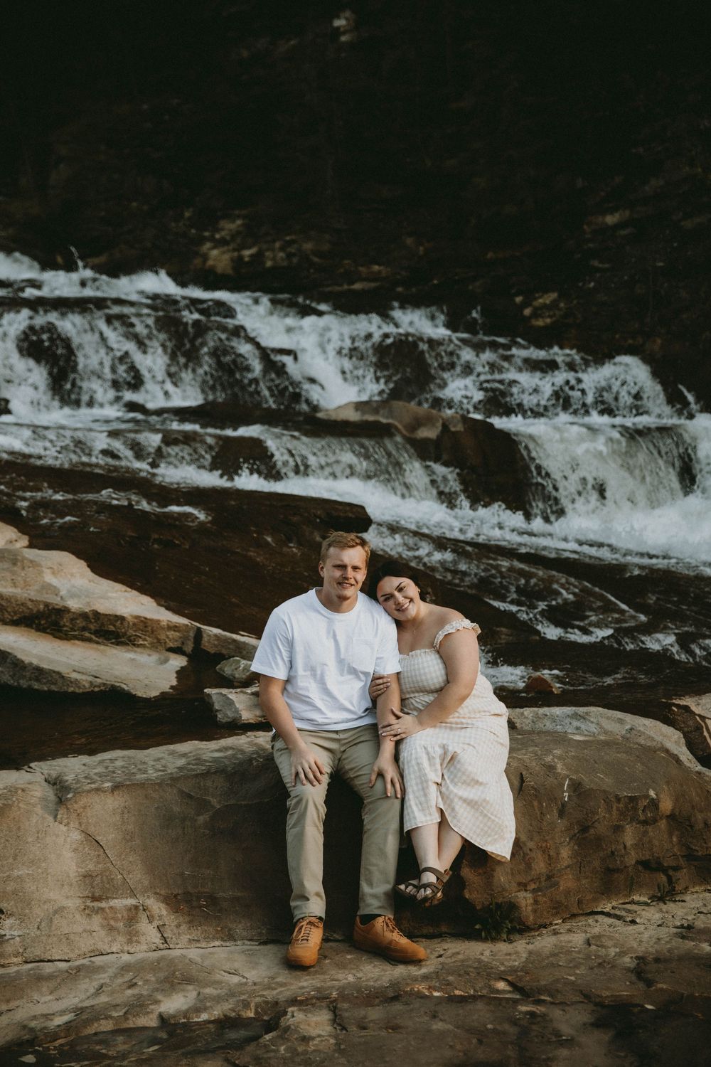 Couple sitting on rocks smiling in front of a cascading waterfall, man in white tee and woman in flowy dress.