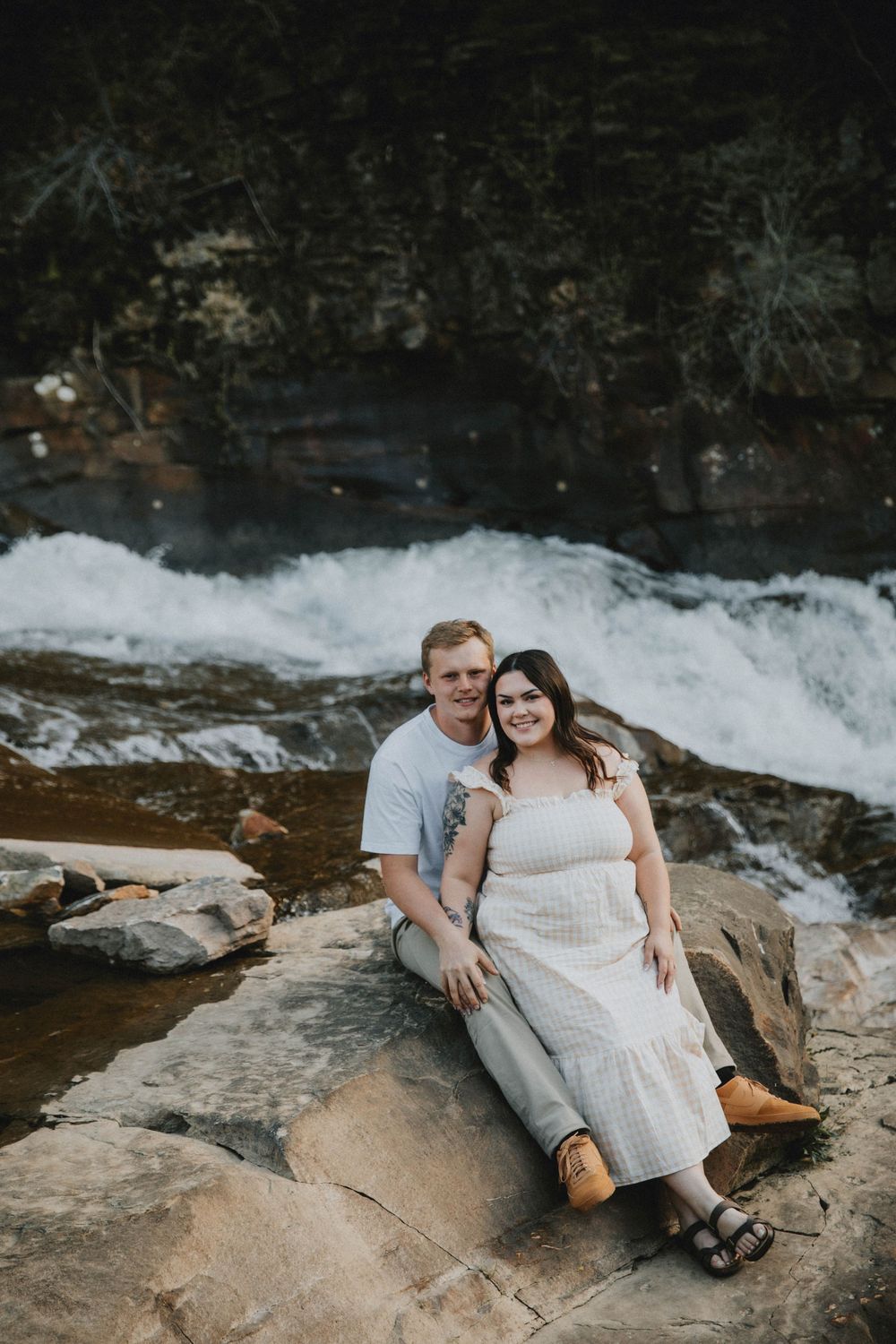 Couple sitting on rocks by a waterfall, woman in white dress and man in white tee, smiling together outdoors. indiana pa photographer