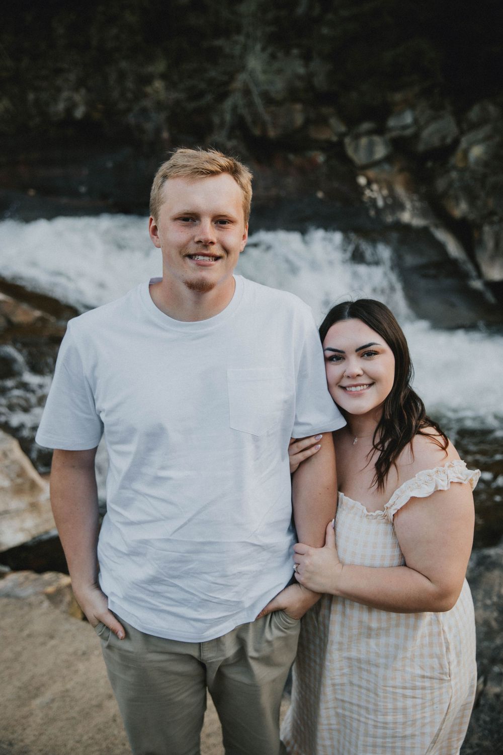 Couple sitting on rocks by a waterfall, woman in white dress and man in white tee, smiling together outdoors. indiana pa photographer