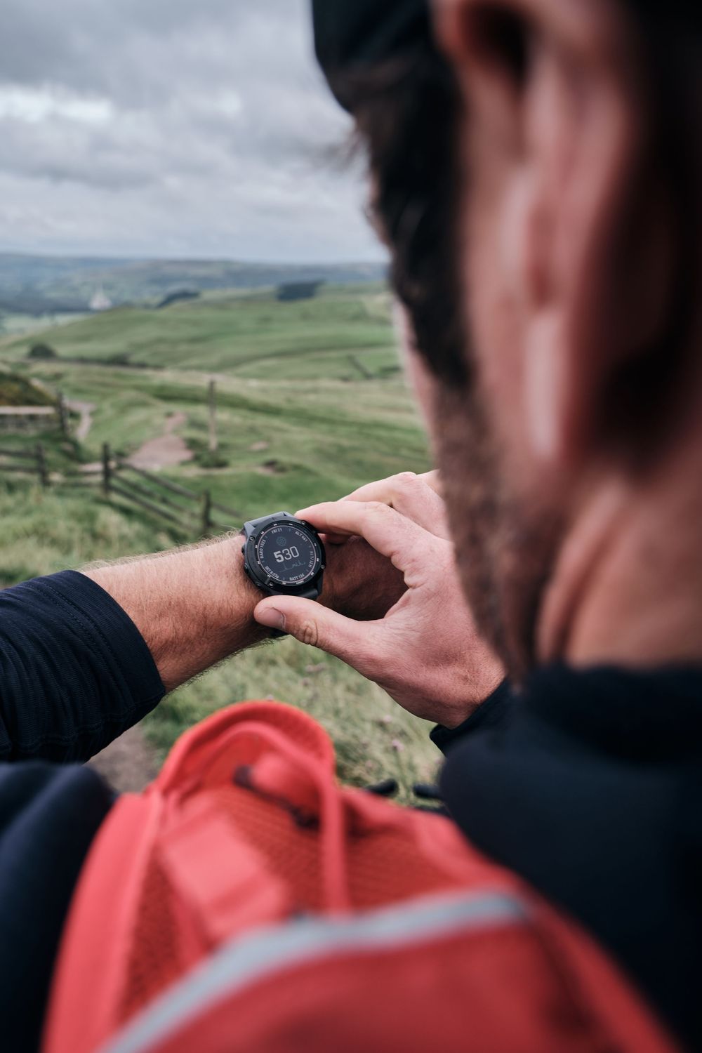 Hiker checks black GPS smartwatch on wrist with green rolling hills landscape in background.