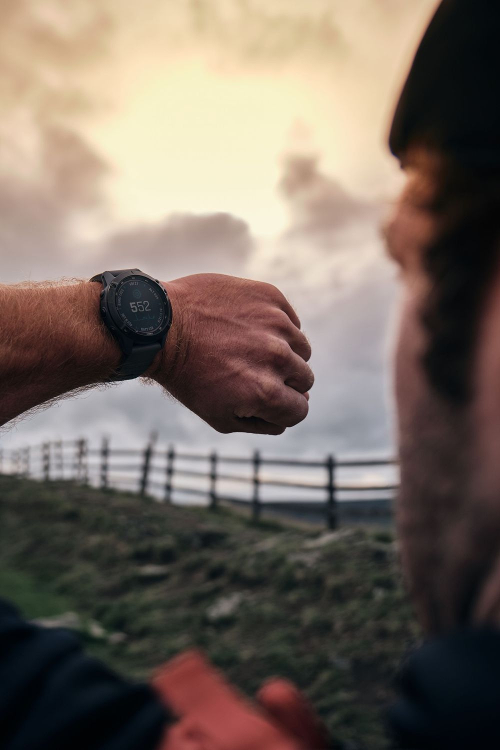 Close-up of hiker's wrist wearing black sports smartwatch against dramatic cloudy sky backdrop.
