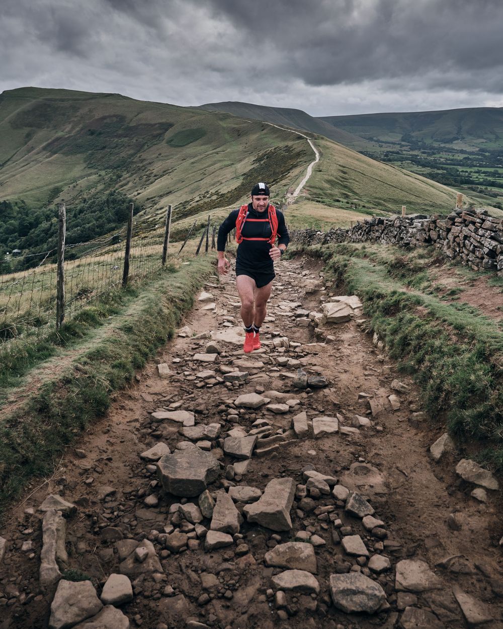 Trail runner in red vest navigating rocky mountain path with rolling green hills and dramatic cloudy sky in background.
