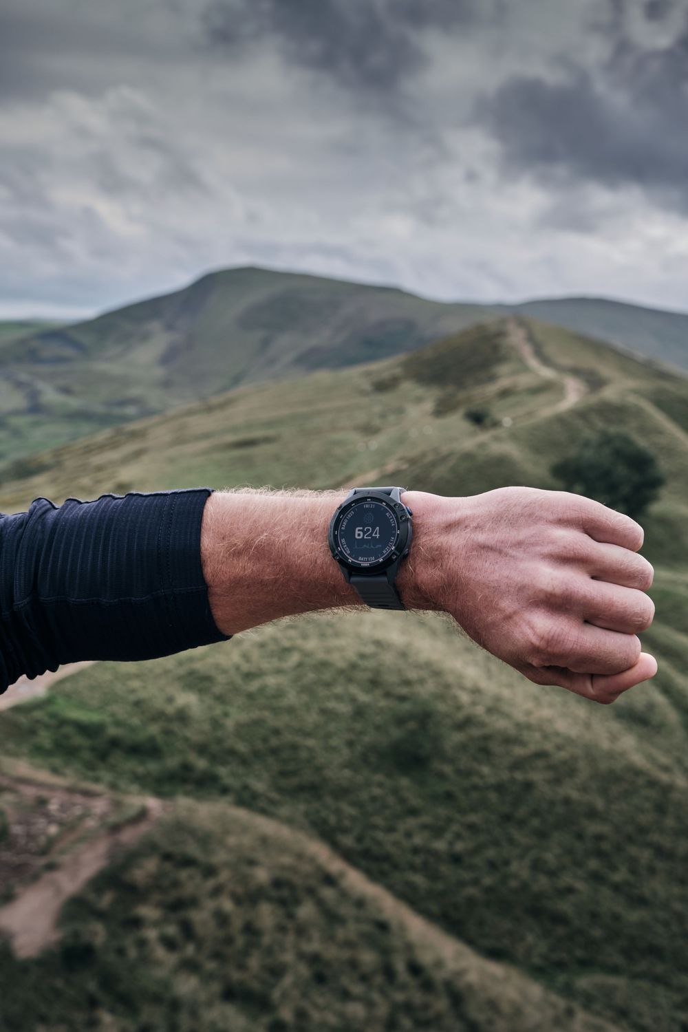 Hiker wearing a black GPS smartwatch on wrist with green rolling hills and cloudy sky in background.