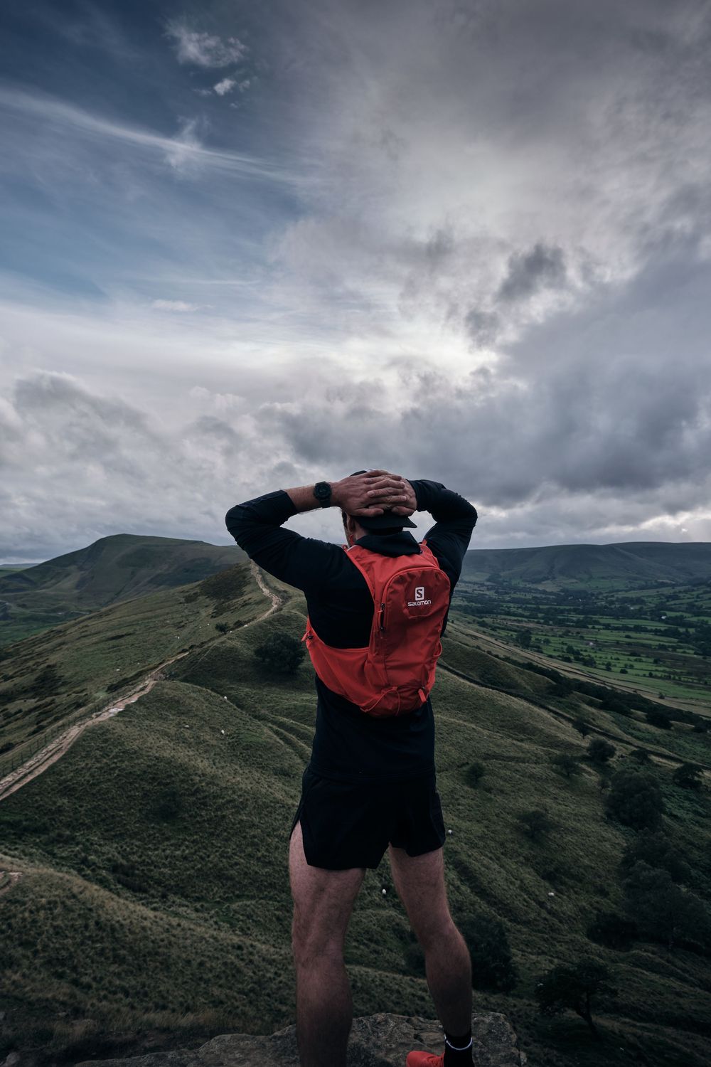 Hiker with red Salomon backpack stands on mountain summit overlooking green rolling hills under dramatic cloudy sky.