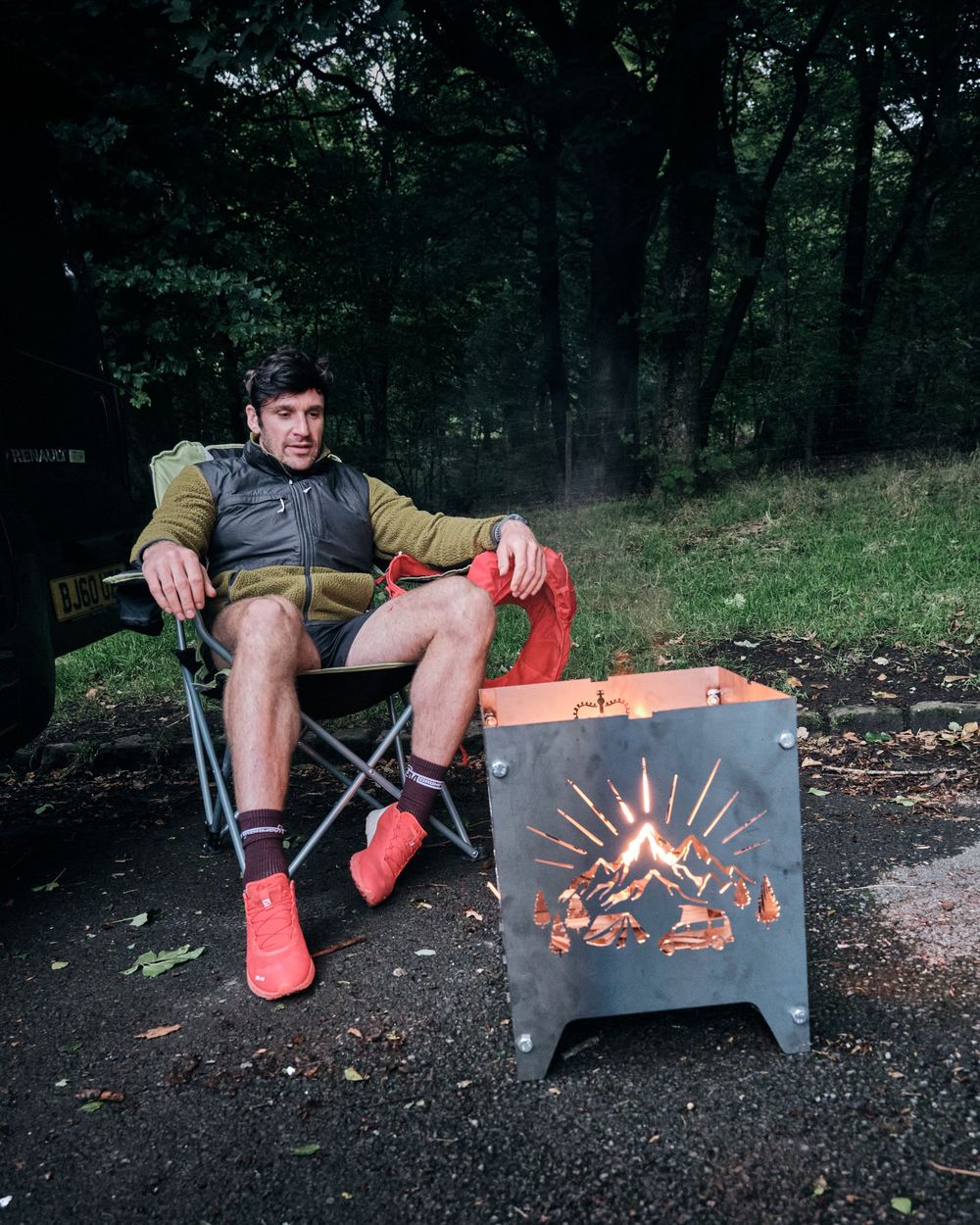 Man relaxing in camping chair beside a decorative metal fire pit with glowing flames in a forest setting.
