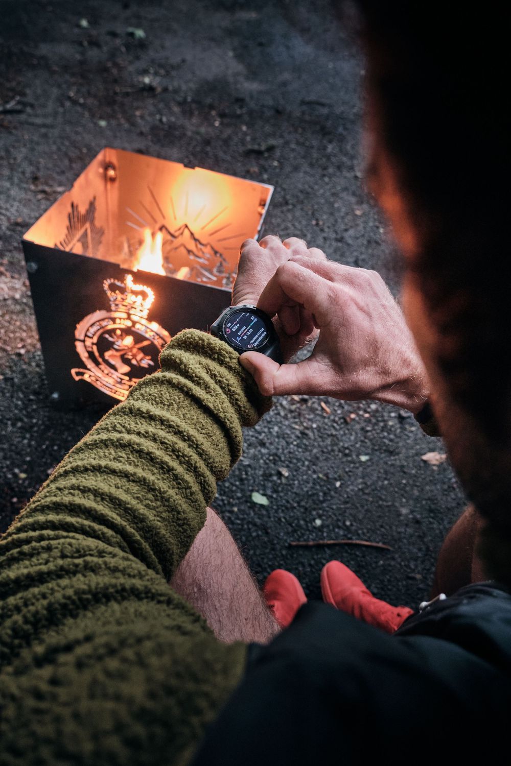Man in green fleece checks smartwatch beside a glowing metal fire box with royal crest emblem outdoors.