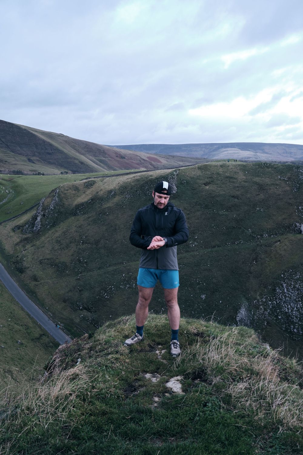 A man in blue shorts and black jacket stands on a hilltop checking his watch with rolling green hills behind him.