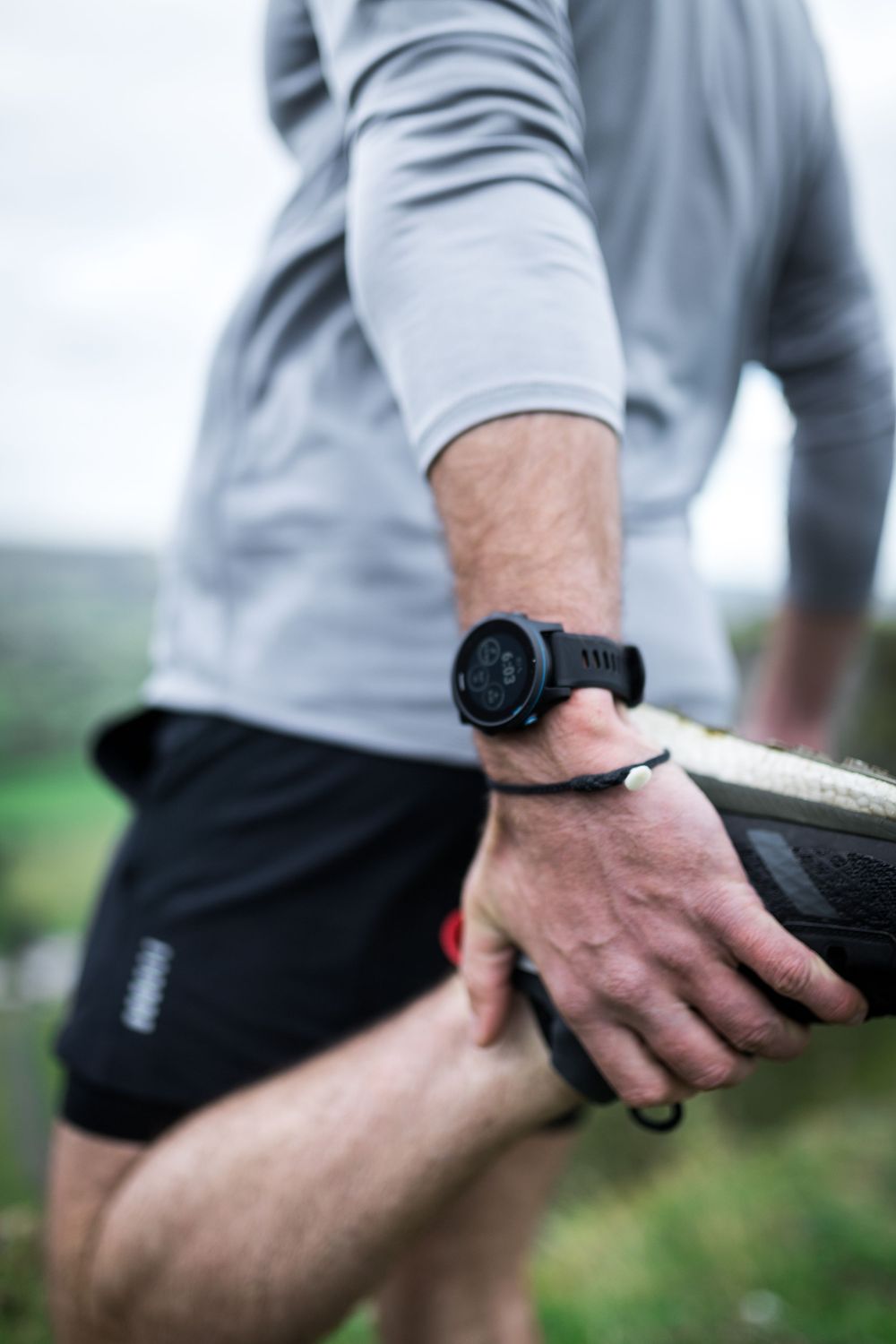 Man in gray shirt and black shorts stretching leg outdoors, wearing a black GPS sports watch on wrist.