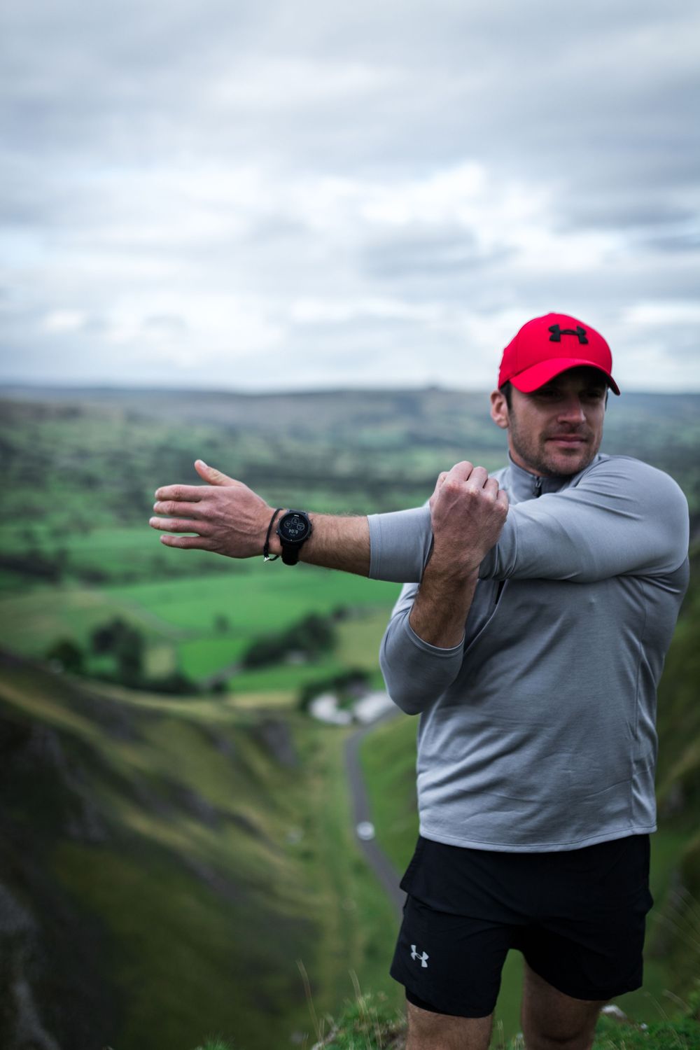 Man in red cap and gray shirt stretching arms on hilltop with green valley landscape behind him.