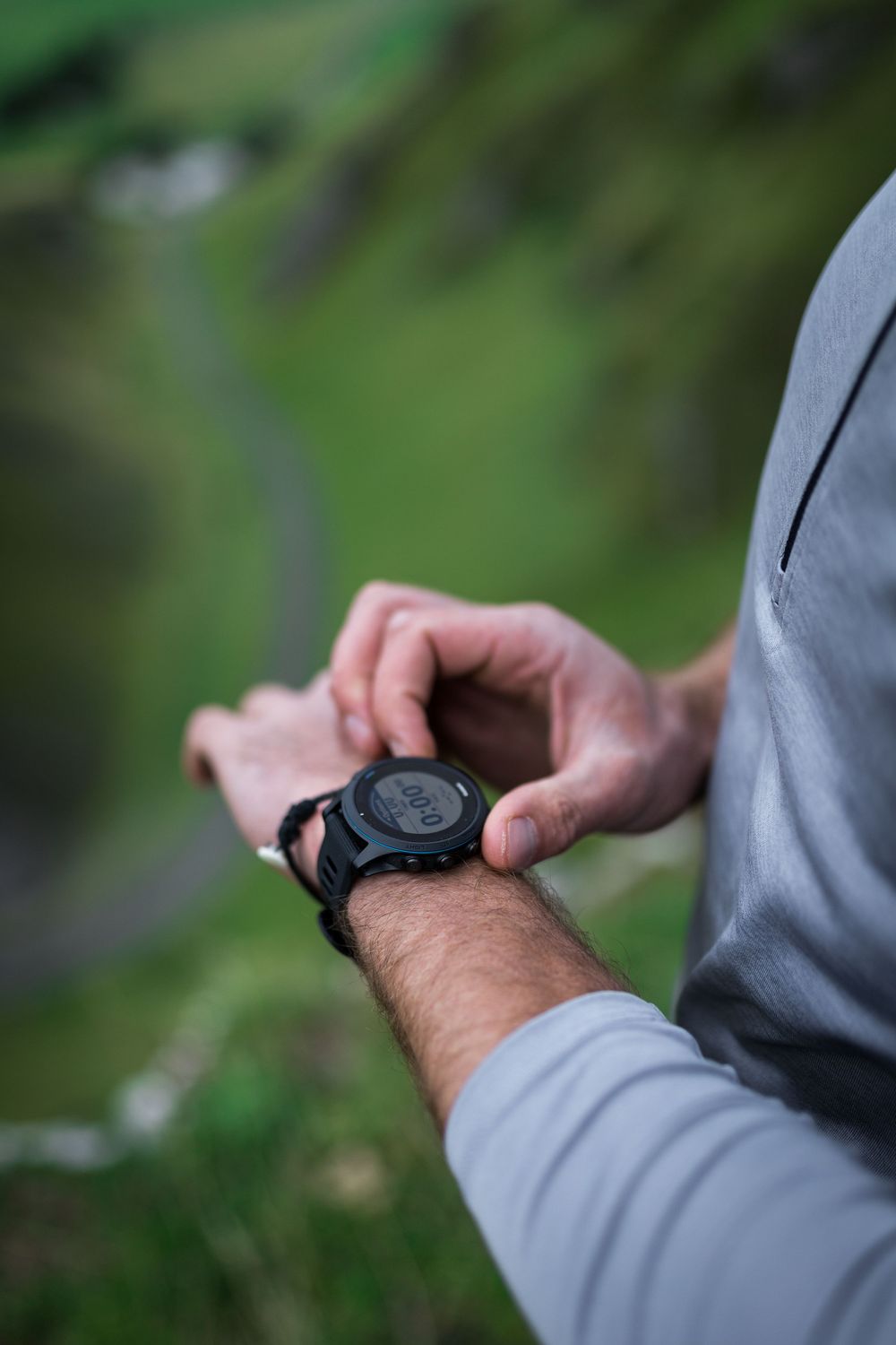 Man checking a black GPS smartwatch outdoors with green mountain landscape in the background.