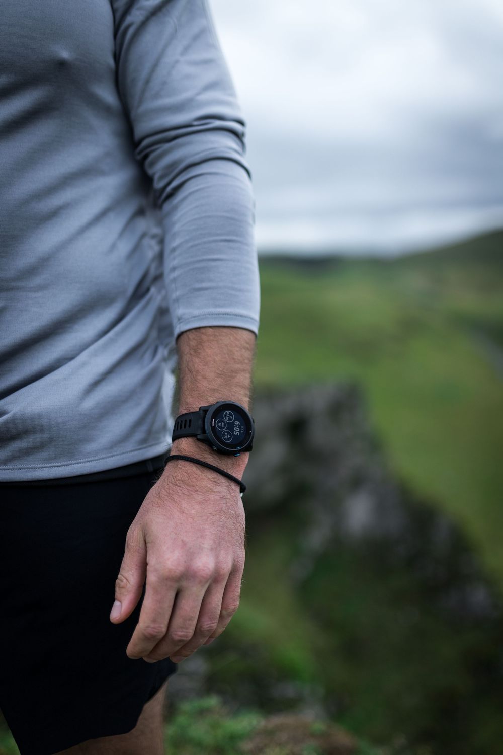 Man wearing a black smartwatch and gray long-sleeve shirt standing outdoors with green hills in the background.