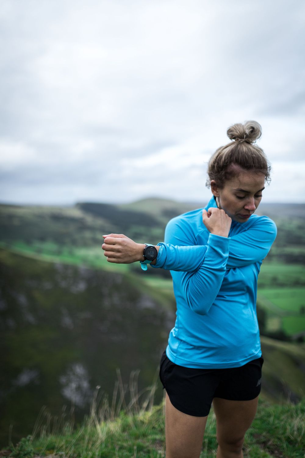 Woman in blue top stretching arms on hilltop with scenic green valley and cloudy sky behind her.