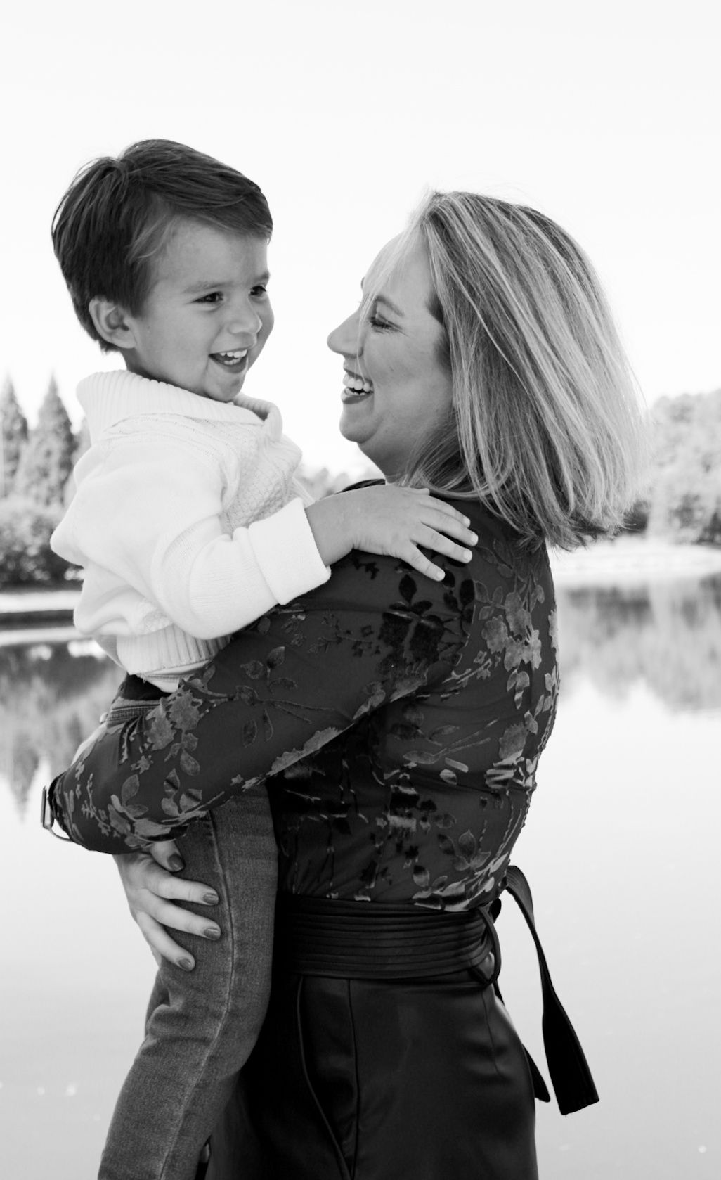 Black and white portrait of smiling blonde woman holding laughing young boy near reflective lake.