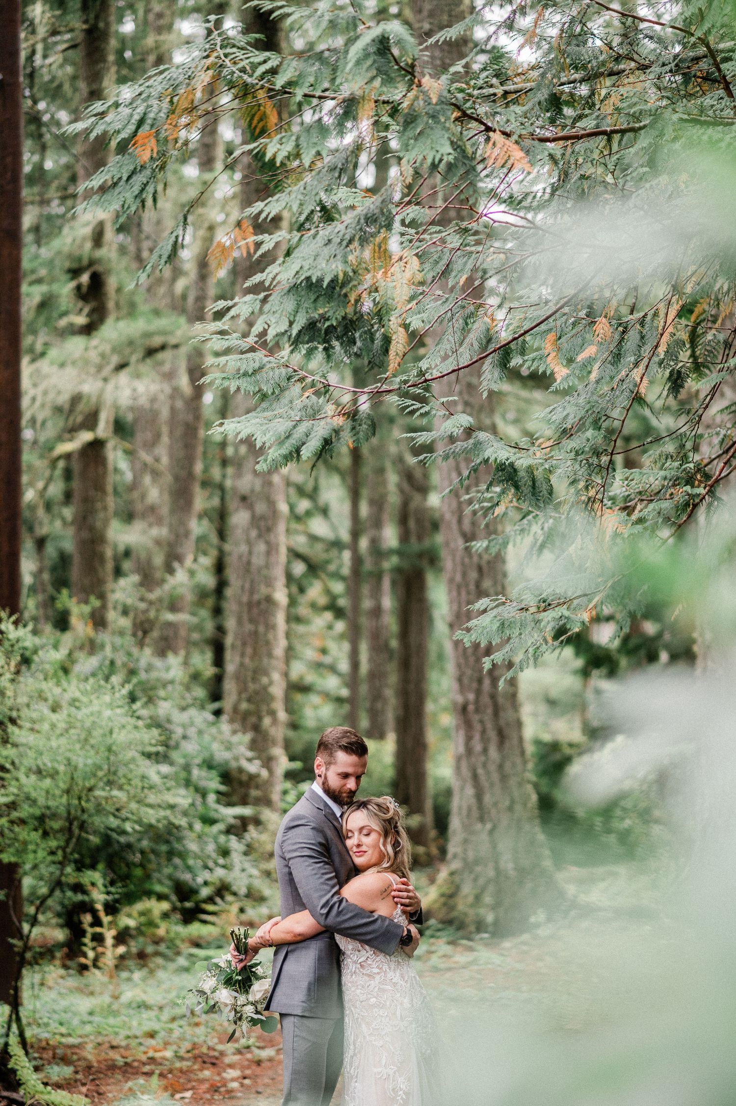 Bride and groom embracing in a lush forest setting, surrounded by tall evergreen trees on their wedding day.