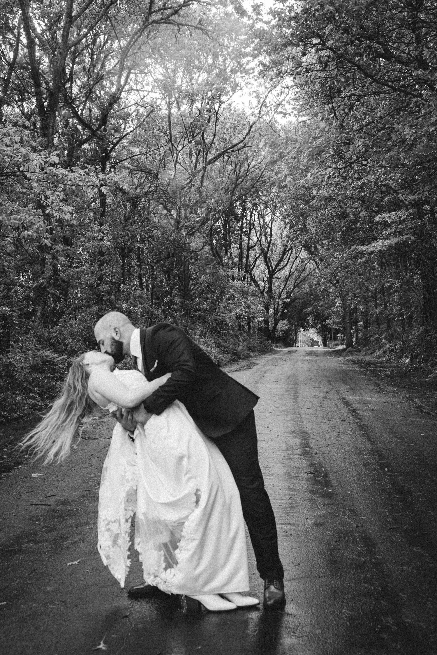 Black and white photo of a bride and groom sharing a romantic dip kiss on a tree-lined road on their wedding day.